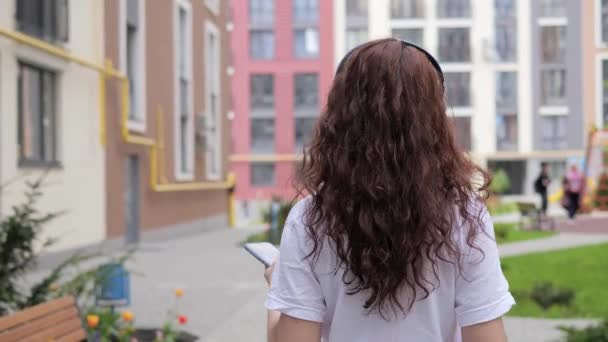 Attractive young curly hair woman with coffee listening to music in wireless headphone using mobile smartphone new app in urban street turning around looking posing to camera walking in the city. — Vídeos de Stock