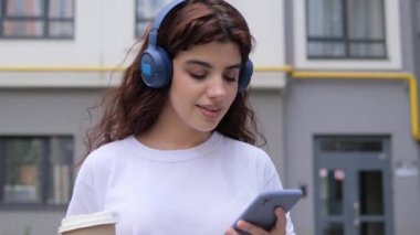 Güzel kıvırcık saçlı kadın, elinde kahveyle kablosuz kulaklıkla müzik dinliyor. Şehirde yürürken cep telefonu uygulaması kullanıyor. Gülümseyen genç bayan hipster kendini özgür hissediyor..