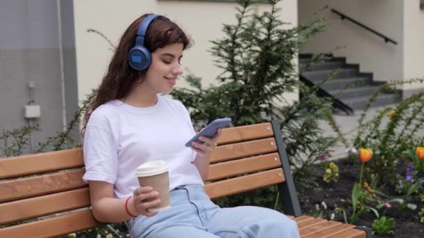 Cheerful curly hair woman in modern wireless headphones relax on bench listening to music. Happy young girl in earphones holding coffee and mobile phone resting outdoors. Enjoy good quality sound. — Vídeos de Stock