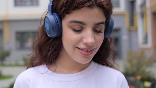 Close up of curly hair woman face in modern wireless headphones relaxing listening to music. Happy young girl in earphones holding coffee and mobile phone resting outdoors. Enjoy good quality sound. — Video Stock