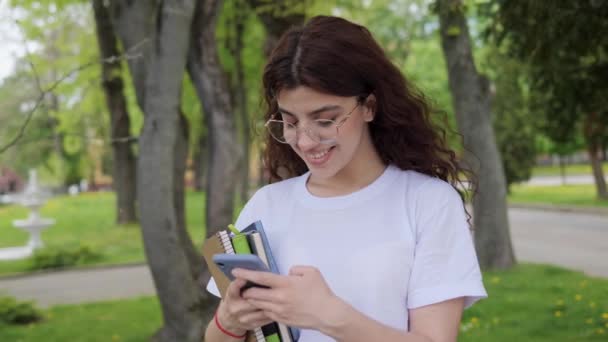 Happy beautiful young curly hair high school girl wearing white t-shirt and glasses messaging on smartphone surfing internet. Student holding notebooks with mobile phone standing in campus outdoors. — Vídeos de Stock