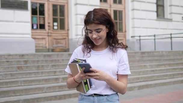 Female curly hair student in white t-shirt with books walking near university using mobile phone in campus outdoor. Beautiful girl surfing social media, surfing internet, tapping screen smartphone. — Vídeos de Stock
