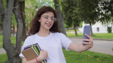 Genç ve çekici bir bayan blogcu video kaydediyor ya da üniversite kampüsü yakınlarındaki bir sokakta görüntülü konuşma yapıyor. Güzel kıvırcık saçlı kız online konferans görüşmesi yapıyor..