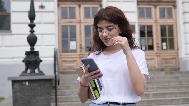 Positive female curly hair student in white t-shirt with books adjusts glasses on the background of college outdoor. Beautiful girl looking to camera. Happy face of young woman with healthy smile. — Vídeos de Stock