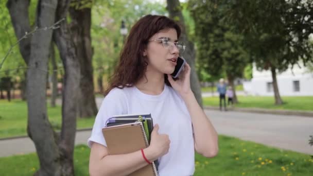 Portrait of curly hair smiling female student talking on phone near university building. Happy caucasian college woman in glasses with notebooks having phone call relaxing on campus after classes. — Vídeos de Stock