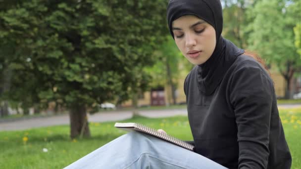 Portrait of young attractive muslim woman in hijab working with laptop reading notes in the park. Charming female student in traditional asian clothes is sitting on the grass near university campus. — Stockvideo