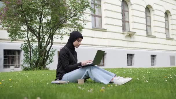 Beautiful muslim arab islamic student girl in hijab studies outside. Woman freelancer user uses laptop sitting on green grass in park near university working online studying remotely chatting outdoor. — Wideo stockowe