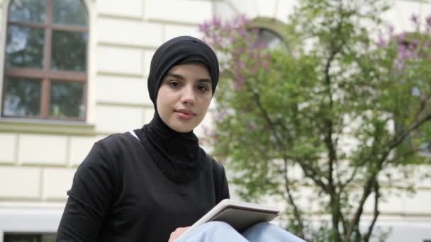 Portrait of a beautiful muslim arab islamic student girl in hijab sitting on a grass near the college. Woman freelancer user uses laptop holding books working online studying remotely outdoor. — Stockvideo