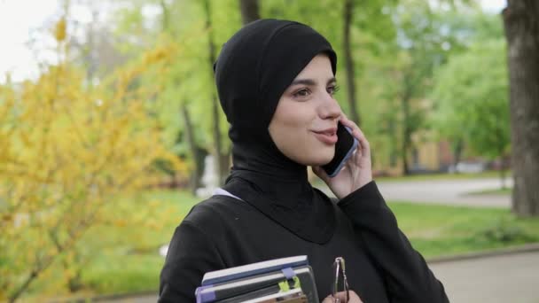 Young Arabic beautiful girl in hijab talking by mobile phone holding textbooks, glasses having nice appearance good mood. Pretty muslim female student holding books, standing in the park near academy. — Vídeos de Stock
