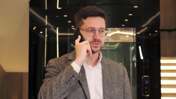 Smiling business man it programmer using smartphone talking on cell phone in modern hallway. Confident professional manager web designer consulting client about online project making business call. — 图库视频影像