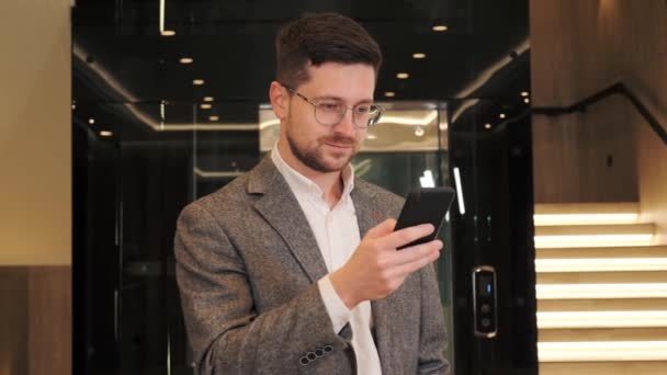 Serious businessman holding smartphone typing message standing near modern elevator. Smart manager ceo using cell phone mobile apps. Digital technology app solutions for business corporate development — Αρχείο Βίντεο