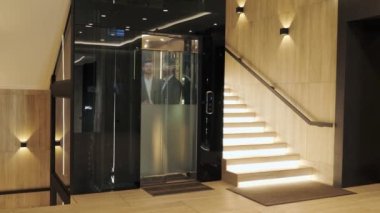 Group of businessmen are moving up in a modern glass elevator. Successful people take the lift to the top of Business Center. Stylish man and woman talking discuss the project inside lift. 