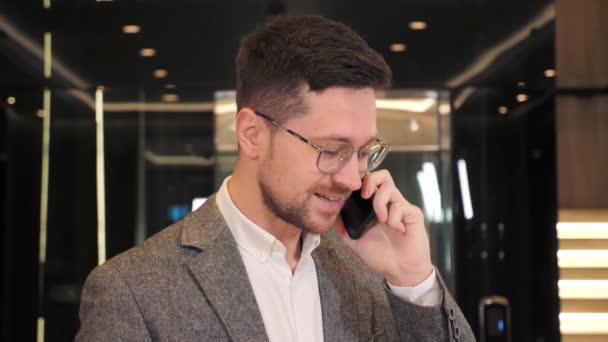 Closeup smiling bearded man in glasses talking on mobile phone in office hall. Close up of cheerful businessman having phone call near elevator. Portrait of happy business man calling phone indoor. — Wideo stockowe
