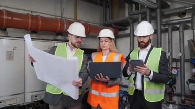 Mechanical engineer holds a plan works on the components and a female chief engineer works on laptop personal computer they discuss heating system in the boiler room on the background of water pipes.