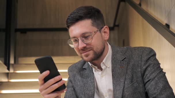 Smart confident man in glasses using smartphone indoors. Businessman mobile technology user working in digital applications gadget searching information online, texting messages in office hall. — Vídeo de stock