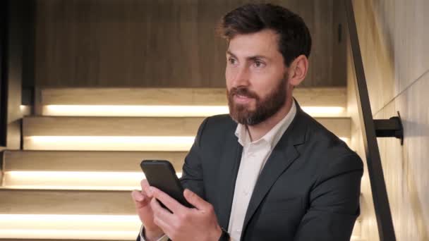 Sorrindo homem focado usando aplicativo smartphone dentro de casa. Empresário usuário de tecnologia móvel trabalhando em aplicações digitais gadget procurando informações on-line, mensagens de texto nas escadas no salão de escritório. — Vídeo de Stock