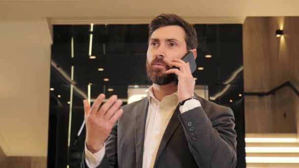 Portre yakışıklı adam iş merkezinde telefon görüşmesi yapıyor. Modern cam asansörün yanındaki koridorda akıllı telefondan konuşan bir iş adamı. İçeriden telefon var. Modern aygıt kullanan bir ofis çalışanı. — Stok video