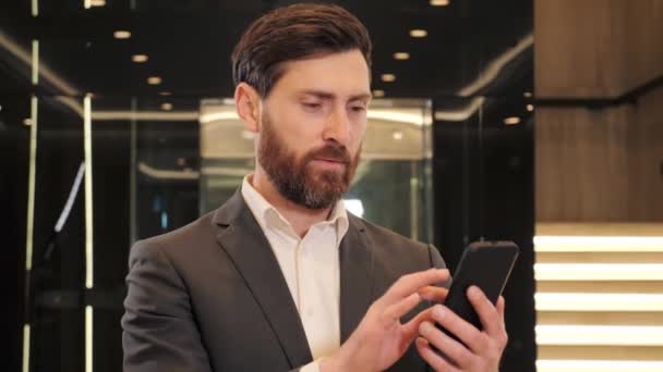 Excited happy business man celebrates victory looking on smart phone, enjoying jackpot in mobile applications. Businessman reading good news on mobile phone in modern hall. Male celebrating victory. — 图库视频影像