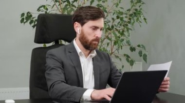 Focused businessman working on laptop holding documents in office. Male professional typing on laptop keyboard at office workplace. Portrait of confident business man makes reporting indoors.