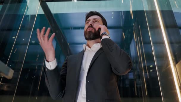 Uomo d'affari di successo carismatico in un vestito cavalcare ascensore di vetro nel moderno centro commerciale. Bell'uomo serio che parla sul cellulare con una chiamata d'affari in ascensore. Leader maschile utilizzando Smartphone. — Video Stock