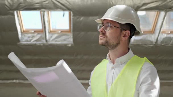 Concentrated young bearded male engineer in white shirt, protective hardhat, glasses standing in unfinished building with blueprints plan and looking away thoughtfully. Thoughtful architect designer. — Vídeo de Stock