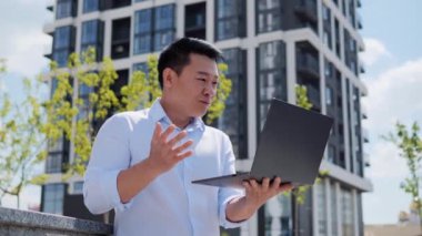 Asyalı mutlu iş adamının portresi dışarıda dizüstü bilgisayardan iyi haberler okuyor. Koreli adam dışarıda başarıyı seviyor. Şaşkın Adam modern binanın yakınındaki zaferi kutluyor. Kazanan ya da kazanan, spor bahisleri