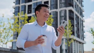 Yakışıklı, gülümseyen Asyalı, genç, yetişkin girişimci iş adamı cep telefonuyla internette sosyal medyada geziniyor ve güzel, modern inşaat sektörünün yanında duruyor. Başarılı Çinli erkek.