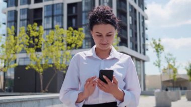 Modern Mobil Aygıtını kullanırken Modern Bina 'nın yanındaki kasabada duran Güzel Yetişkin İş Kadını. Kendinden emin ve meşgul görünüyorsun. Haberleri ve postaları okuyorum. Güzel görünüş, güneşli bir gün.