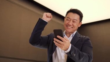 Mutlu Asyalı işadamının portresi cep telefonunda başarılar. Yakın plan sevinçli Çinli adam telefondan iyi haberler okurken elini kaldırıyor. Şaşkın erkek telefonda zaferi kutluyor.