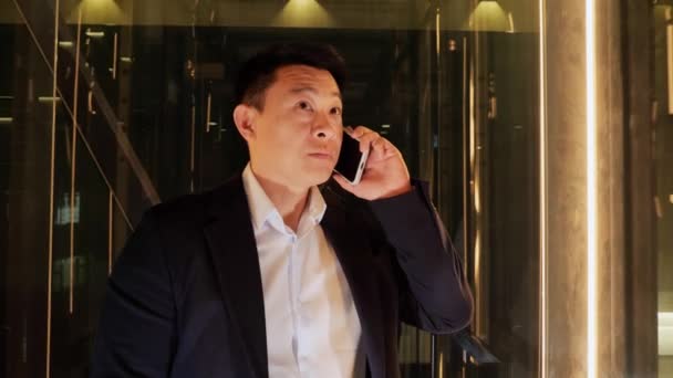 一位亚洲英俊的商人 一位年轻的高层经理 坐在一幢办公大楼的全景电梯里 一边用手机聊天 中国男人看上去专注而自信 成功的商人 — 图库视频影像