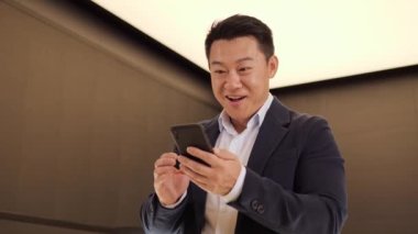 Mutlu, şanslı, takım elbiseli, cep telefonlu, komik suratlı, heyecanlı iş adamı akıllı telefonuna bakıp iyi haber e-postalarını okuyan, duygusal yaşam tarzı konsepti olan mutlu, heyecanlı genç Çinli bir adam.