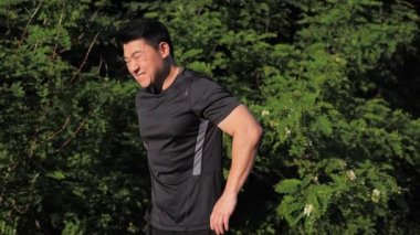 Duran genç Asyalı koşucu, yeşil ağaçların arka planında egzersiz yaparken sırtında acı hissediyor. Sporcunun omuz ağrısı, sakatlığı var. Spor koşucusu gün batımında koştuktan sonra omuz ağrısı çekiyor.