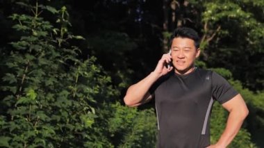 Güneş ışığı tebessümünde cep telefonu standında konuşan yakışıklı Asyalı adam. Erkek koşucu dışarıda sağlıklı spor yapıyor. Spor sonuçlarını paylaşan bir arkadaşla cep telefonu konuşması. Koşucu çevrimiçi iletişim