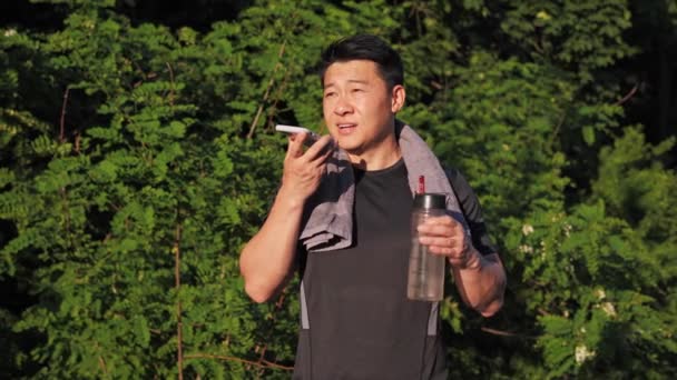 屋外を走った後 スピーカーのスポーツ結果を共有する上での音声メッセージを記録夏の公園に立って彼の肩の上にタオルで快適なアジアの男 ワークアウトを終え 新鮮な空気を楽しむ — ストック動画