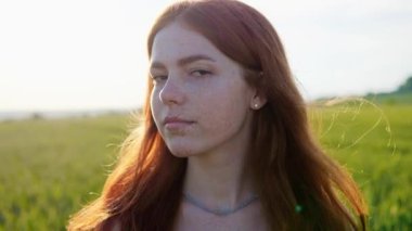 Gün batımında tarlada güzel kızıl saçlı bir kızın portresi. Yeşil buğday tarlasında kameraya bakan güzel kızıl kadın. Uzun saçlı, çilli, kahverengi gözlü güzel bir kadın yüzü.