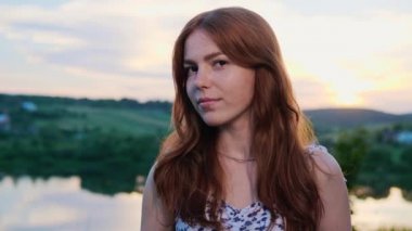 Güneşin batışında gölün arka planında duran kızıl saçlı güzel mutlu kızıl kadın kameraya bakıyor. Dışarıda gülümseyen sevimli bayan. Duygular. - İnsanlar. Portre