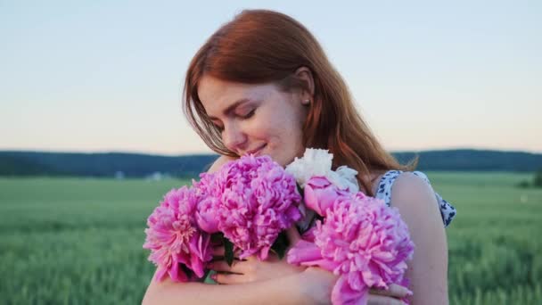 可爱美丽的红头发少女带着雀斑的特写 女人牵着粉红花朵望着夕阳西下麦田背景的相机 — 图库视频影像