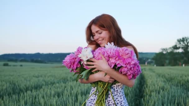 幸せな美しい赤髪の若いジンジャーガールとそばかす ピンクの花の牡丹を持つ女性は 外の夕日で緑の麦畑の背景に魅力的な笑顔を楽しんでいます — ストック動画