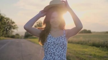 Yazlık elbise giymiş çilli kızıl saçlı kız gün batımında yolda yürüyor. Güzel kızıl kadın hasır bir şapka takıyor ve dışarıda şakacı bir şekilde gülümsüyor. Güzel doğa, manzara, yaz