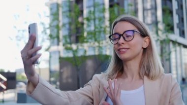 Gözlüklü, gülümseyen genç bir iş kadını iş yerinin önünde selfie video görüşmesi yapıyor. Modern ofis binasının yakınlarında video blogu çeken bir iş adamı portresi. 