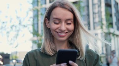 Modern ofis binasının yakınında akıllı telefon kullanarak gülümseyen mavi gözlü genç bir iş kadını. Yeşil tişörtlü güzel bir kız sokakta dikilirken sosyal ağlarda flört uygulaması kuruyor.