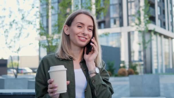 市内で素敵な日に友人と電話を持っている魅力的な積極的な服装のブロンドのビジネスマンの女性 コーヒーを持っている白人女性フリーランスオフィスの外に立つ — ストック動画