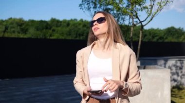 Güneş gözlüklü genç sarışın iş kadınının portresi cep telefonuyla yürüyor göğsünde ağrı, açık havada derin nefes alıyor.