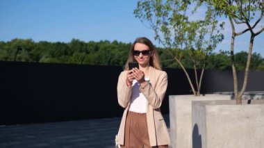 Şehir sokağında durmuş, son moda güneş gözlüğü takan gülümseyen kadın cep telefonunu kullanıyor. Kahverengi ceketli güzel bir yaz kadını internet, sosyal medya, flört uygulaması ararken akıllı telefona bakıyor.