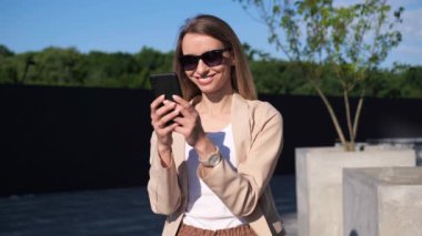 Güneş gözlüklü, dışarıda cep telefonu kullanarak duran sevimli çekici kadın gün ışığında mutlu bir gülümseme hisset. Gezgin kadın akıllı telefon internet, sosyal ağlar, flört uygulamaları kullanıyor.