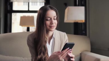 Otel odasında cep telefonu kullanan gülümseyen bir kadın. İş kadını mesaj iletmek için çalışıyor. Akıllı telefonları tarıyor, internette geziniyor. Mutlu kadın iş otelinde cep telefonuyla koltukta oturuyor.