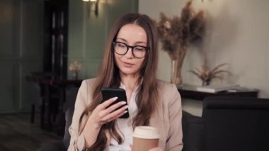 Gözlüklü bir iş kadını otel lobisinde elinde kahve tutan cep telefonu uygulaması kullanıyor. İş yerinde çay içen gülümseyen kız. Kafede internette sörf yapan mutlu iş kadınlarını kapatın.