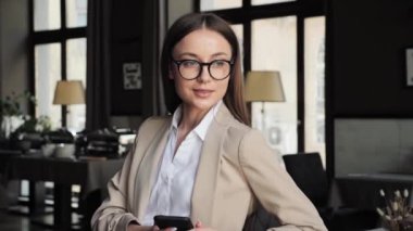 Elegant happy young pretty business woman wearing formal outfit in eyeglasses sitting at table. Good looking caucasian girl holding smartphone in hands, chatting working from restaurant indoor