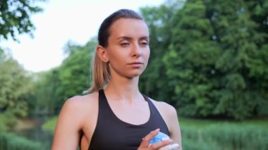 Atletik sporcu kadın gün batımında şehir parkında spor şişesinden su içiyor ve gülümseyen kameraya bakıyor. Yeşil doğa antremanından sonra kadın susuzluğunu giderir. Kadın enerji içeceği sulanıyor.