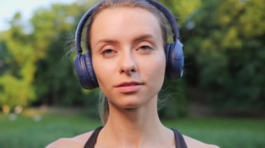 Neşeli, pozitif, güzel bayan sporcu mavi kulaklıkla müzik dinlerken dışarıda güneş batarken kardiyo çalışmasından sonra kameraya poz veriyor. Motivasyon. Özgürlük için. Keyif